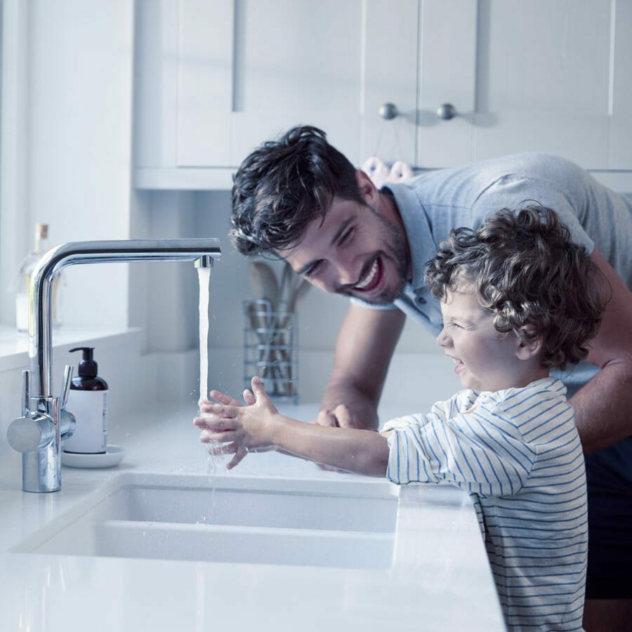 Man and child laughing while child washes his hands under sink.